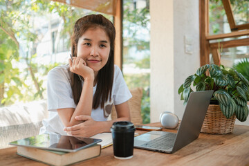 Young woman sitting at a wooden desk with a laptop, tablet, and coffee, enjoying a bright and cozy home workspace.