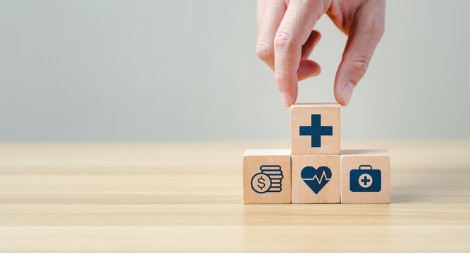 Hand placing a wooden block with a medical cross symbol on top of other blocks featuring healthcare, finance, and insurance icons, representing health services, medical support, and financial coverage
