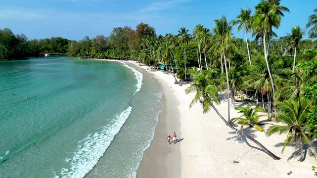 Couple strollers enjoy a romantic beach walk on Koh Kood Island, as soft waves lap against the shore and palm trees sway gently. A tranquil escape in Thailands hidden paradise awaits.