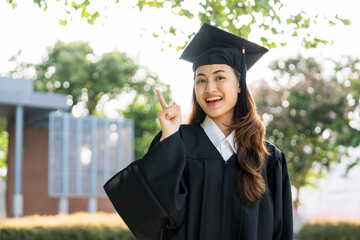 Happy asian woman in graduation costume with cap standing outdoor public park. College degree...