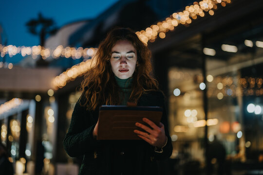 A young woman engaged with a tablet device outdoors, illuminated by evening ambient city lights.