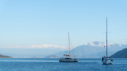 Fototapeta premium Two sailboats float on the Mediterranean sea at Fethiye, Turkey, with a stunning backdrop of snow-capped mountains and a clear blue sky, creating a serene maritime scene.