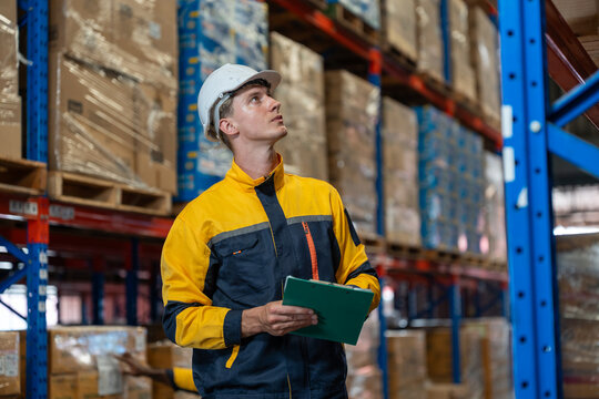 Man warehouse worker wearing uniform and helmet safety standing with clipboard for checking stock products on shelves in warehouse. Logistics, Distribution Center concept