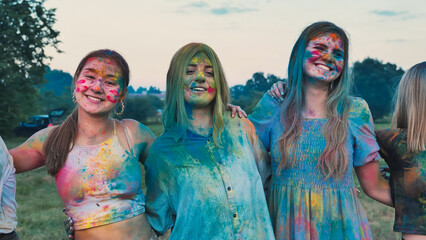 Three school friends embrace and smile, covered in colorful holi powder paint, enjoying a vibrant...