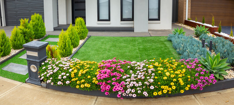 Modern front yard features neatly maintained artificial turf or synthetic grass, bordered by flower beds. A low-maintenance landscape with a small garden and a well-organized entrance to a house.