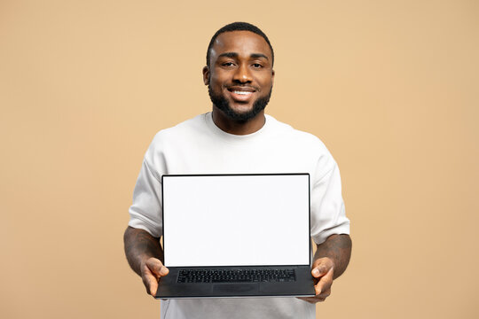 Smiling African American man showing laptop with blank screen on beige background