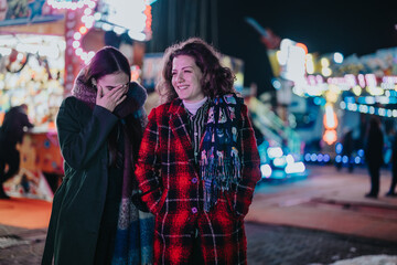 Fototapeta premium Two laughing friends warmly dressed are at a night carnival, enjoying the winter atmosphere and colorful lights.