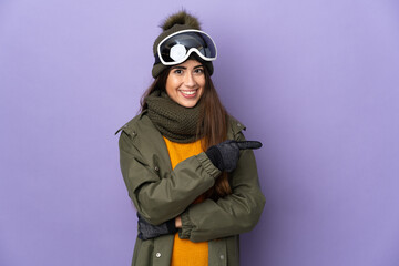 Skier caucasian girl with snowboarding glasses isolated on purple background pointing finger to the...