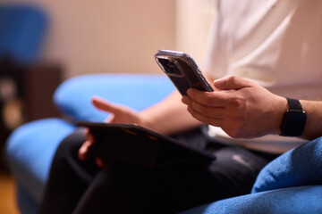 A person is seen sitting comfortably on a blue couch while using both a smartphone and a tablet