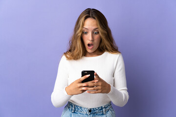 Fototapeta premium Young blonde woman isolated on purple background looking at the camera while using the mobile with surprised expression