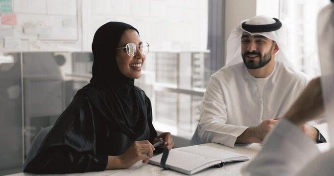 Smiling woman in abaya engaged in conversation with two male teammates dressed in kandora, participate in professional business meeting in office, planning strategy, cooperate, share ideas or opinions