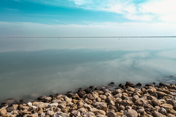 A still horizon where sky meets water, mirrored in perfect calm. Gentle waves kiss the rocky shore...
