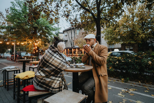 A couple sits at an outdoor cafe, wrapped in blankets, enjoying hot drinks. Autumn leaves and cozy atmosphere create a warm and picturesque setting. Perfect image for themes of love and warmth.