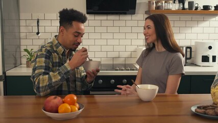 Multicultural couple having breakfast in the kitchen. Man eats cereal while woman talks and smiles during a morning conversation. - Powered by Adobe