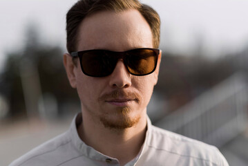 Close-up portrait of a Caucasian redhead young man wearing sunglasses and stylish clothes. The man confidently looks at the camera while walking along the street. Business, career.