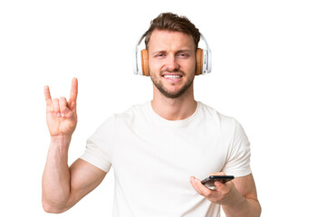 Young handsome caucasian man over isolated chroma key background listening music with a mobile making rock gesture