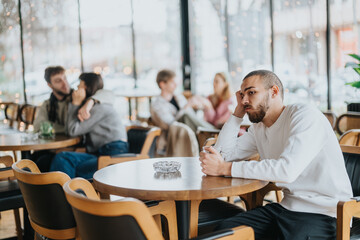 A man sits alone at a table in a bright, cozy cafe. In the background, people socialize and enjoy each others company, highlighting themes of loneliness and human connection.