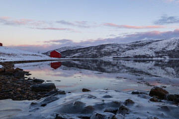 rotes Rorbuer-Haus an einem ruhigen See in Norwegen bei sanftem Abendlicht, der Himmel zeigt ein...
