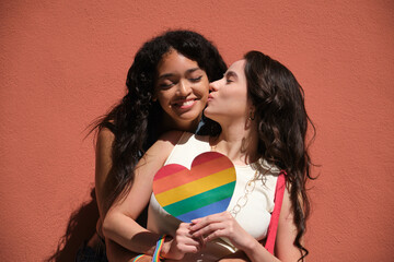 Two young women embracing each other while holding a heart shaped rainbow flag, symbolizing LGBT...