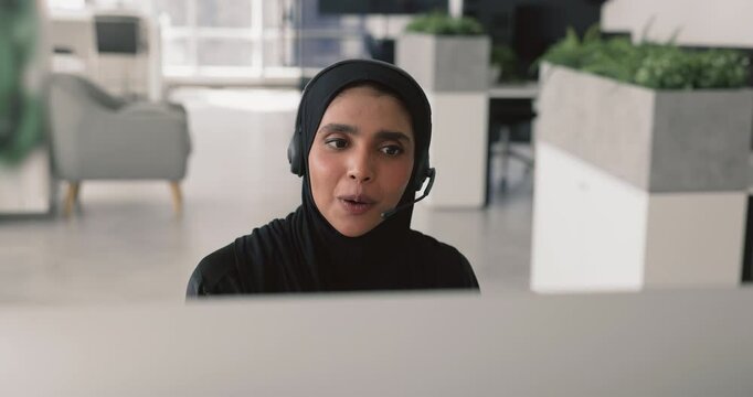 Close up smiling Arabian woman, live chat assistant in black abaya working in office, wear headset provide real-time support through chat, guiding, helps with problem-solving, talks to client remotely