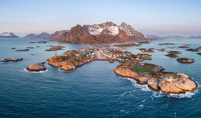 Henningsvær auf den Lofoten in Norwegen bei klarem Himmel am Morgen, die verschneiten Berge und der Fußballplatz auf der Insel von der tief stehenden roten Sonne angestrahlt, eine winterliche Szene