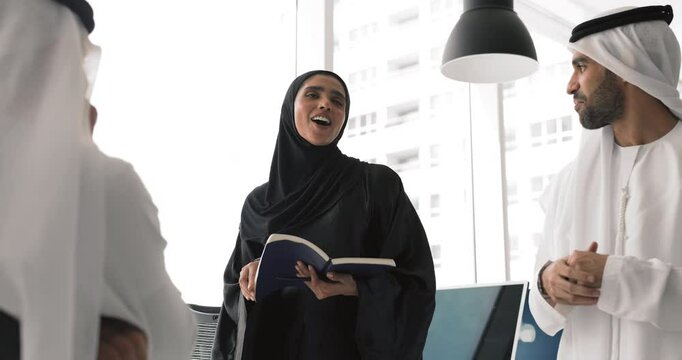 Leadership, empowerment, professional communication. Woman in black abaya holds notebook, speaks with confidence, share ideas, point and solutions with two male colleagues during collaborative meeting