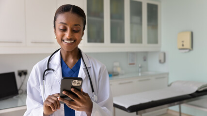 Cheerful young Black physician woman typing on mobile phone in clinic office, smiling, enjoying...