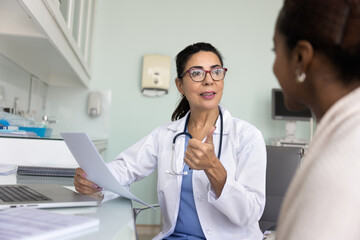 Fototapeta premium Friendly engaged middle aged elder doctor giving consultation to African patient, talking to young woman in office, holding paper medical report, history, giving prescription, explaining treatment