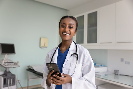 Happy beautiful African doctor woman holding smartphone, posing in hospital examination room, looking at camera, smiling, using online medical professional application on cellphone