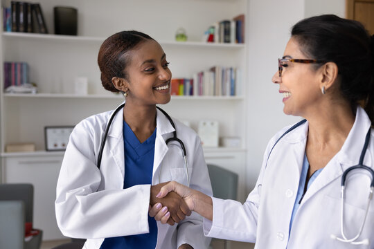 Happy young African American doctor woman shaking hands with senior female colleague in white coat, smiling, laughing, thanking for professional help, advice, support, mentorship, teamwork - Powered by Adobe
