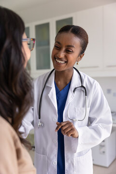 Cheerful sympathetic young Black doctor woman talking to patient on consultation, giving support, appraisal, telling good news after checkup, giving recommendation for healthcare. Vertical shot