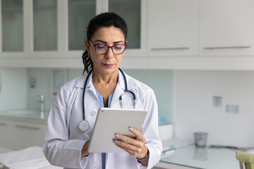 Focused mature physician woman using modern technology on digital tablet in medical examination...