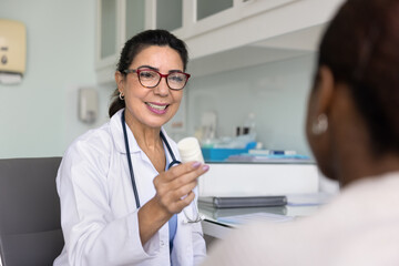 Positive mature Latin doctor woman giving medication to young patient, offering bottle of pills, effective food supplement, painkillers, telling about successful treatment, smiling