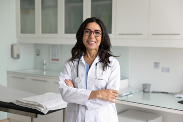 Happy mature female doctor in white uniform coat and glasses posing in hospital examination room,...