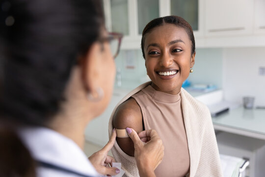 Cheerful young African patient woman visiting doctor for vaccination, talking to physician, nurse sticking plaster on shoulder after injection, smiling, getting vaccine for protection from flu virus