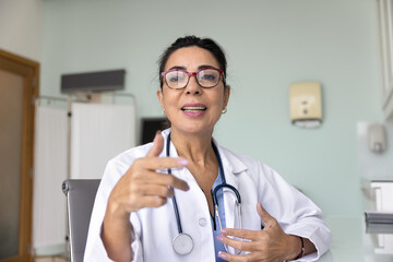 Positive mature Latin practitioner woman giving online consultation, looking at camera, speaking,...