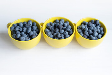 Fresh juicy wild blueberries in three yellow mugs close-up on white table surface, top view, soft focus