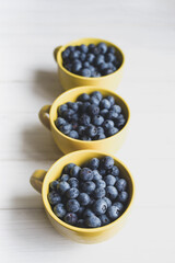 Fresh juicy wild blueberries in three yellow mugs close-up on white table surface, top view, soft focus