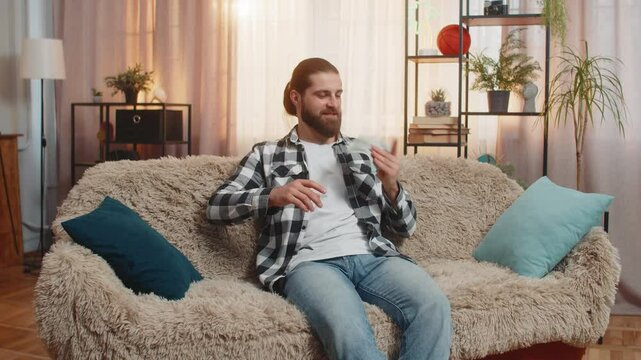 Young man arrives at home with a stack of dollar cash, flinging scatters throwing banknotes upward in celebration. Caucasian guy on sofa carefree spending money, marking financial success playfully