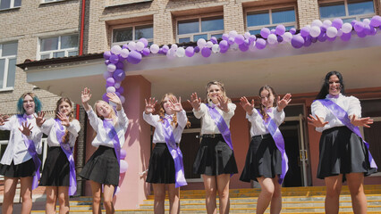 Cheerful female graduates waving goodbye on their last day of school, celebrating the end of the academic year with purple ribbons and balloons © Довидович Михаил