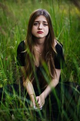 Sad young woman with long brown hair sitting in tall grass, wearing a black dress, embodying feelings of loneliness and melancholy