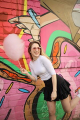 A joyful young woman is holding a stick of cotton candy while posing next to a colorful mural. She expresses happiness in a playful outfit on a sunny day, showcasing urban creativity.