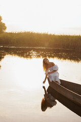A woman in a white dress sits in a boat on a lake at sunset, dipping her hand into the water. Warm...