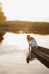 A woman in a white dress sits in a boat on a lake at sunset, dipping her hand into the water. Warm...