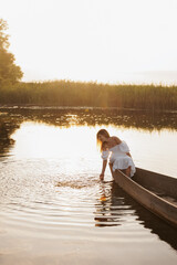 A woman in a white dress sits in a boat on a lake at sunset, dipping her hand into the water. Warm...