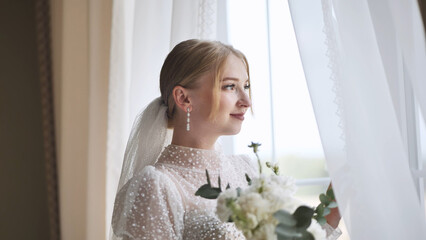 White clad bride holding wedding bouquet, gazing through window with soft, romantic expression