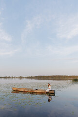 A young woman in a white dress sits on the edge of a wooden boat in the middle of a lake covered with water lilies. Calm water, reflection, and evening light create a dreamy and romantic atmosphere.