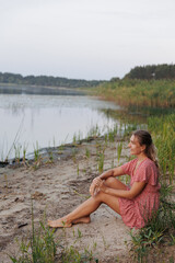Fototapeta premium A woman in a red dress sits barefoot on the lakeshore, surrounded by wild nature. She smiles and enjoys the tranquility, creating an atmosphere of harmony, relaxation, and connection with nature