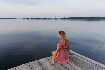 A woman in a light red dress sits barefoot on a wooden pier by the lake, smiling. The evening sky,...
