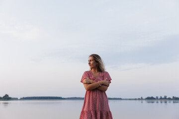 A young woman stands barefoot on a wooden pier by a calm lake. She wears a summer dress and has a...